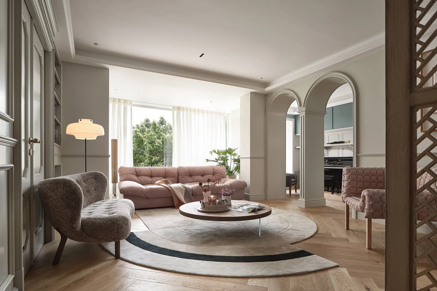 A modern living room featuring a pink sofa, round wooden coffee table, and glass window with greenery outside.