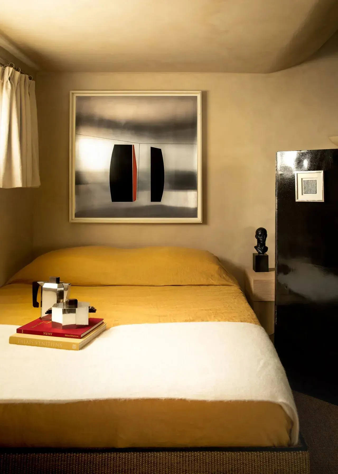 A modern bedroom with a yellow linen bed featuring a painting, a sculpture, and a coffee maker.