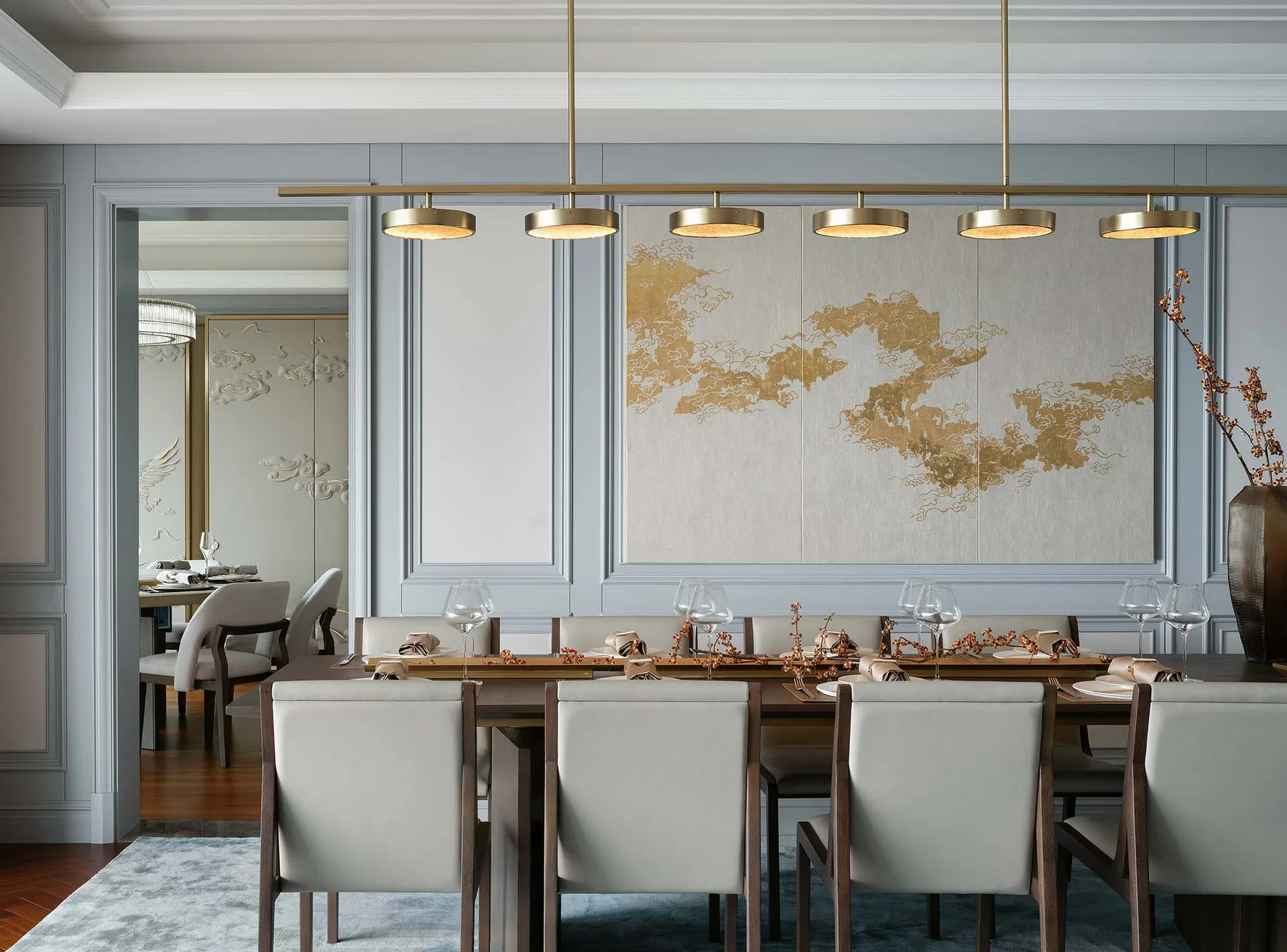 A modern dining room with a large table, chairs, and decorative wall art featuring gold patterns on a light background.