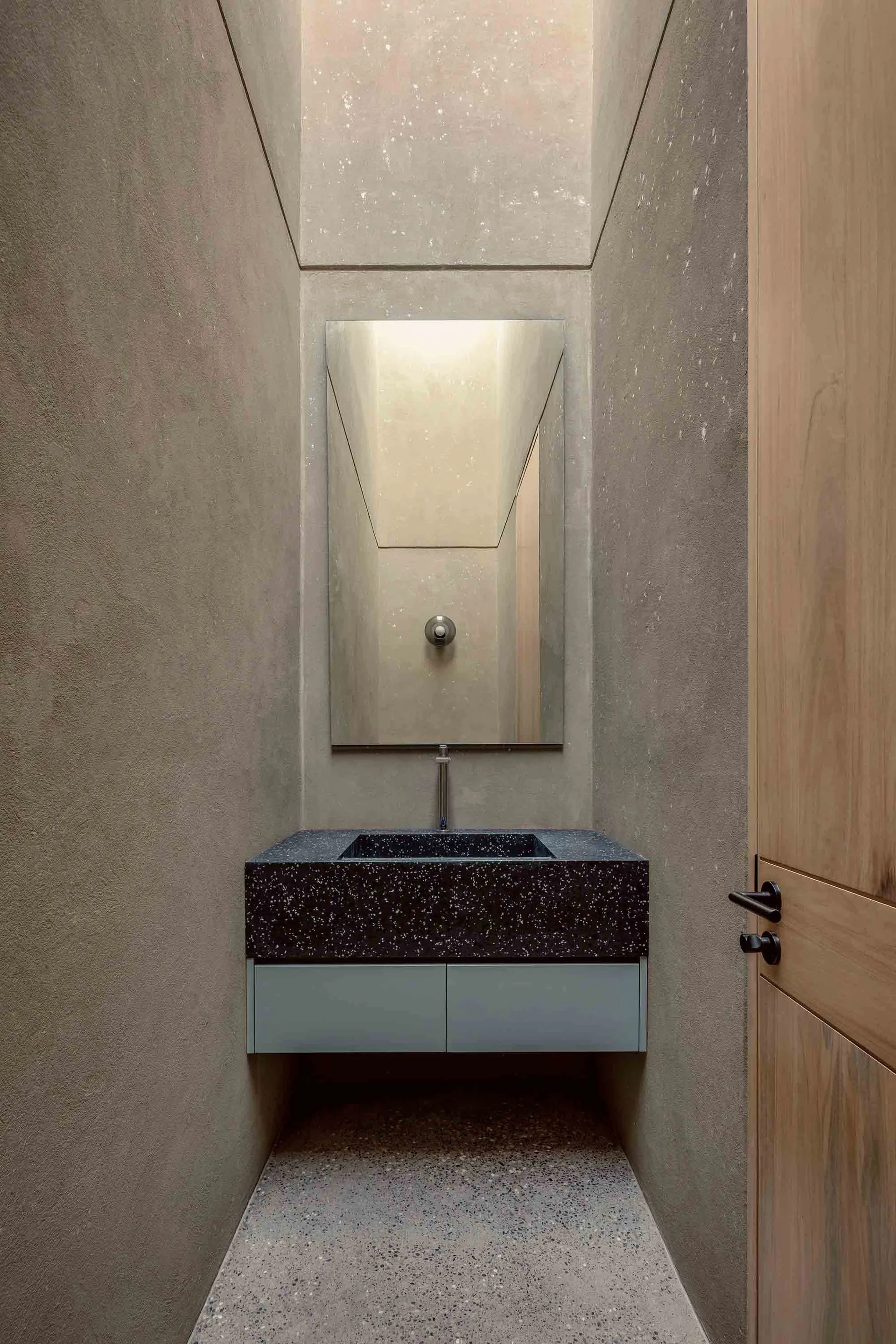 Modern minimalist bathroom featuring a black and white speckled sink counter, a rectangular mirror, and wooden door.