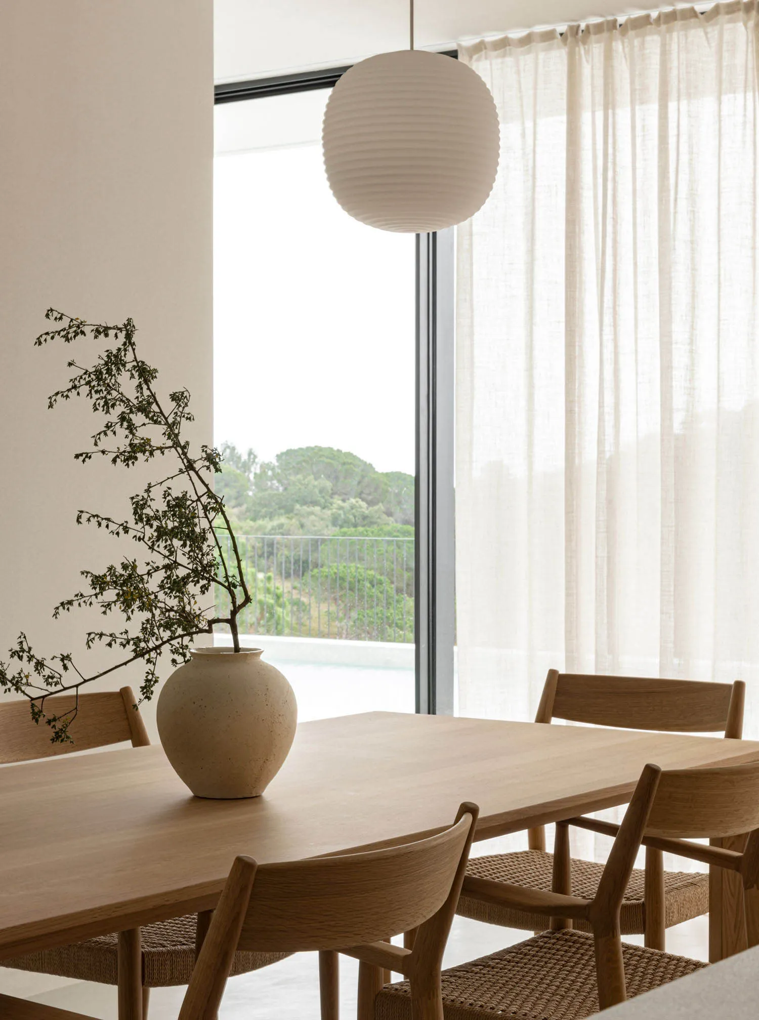 A modern minimalist dining room with a wooden table, chairs, and a vase with plant branches on it, near a large window with w