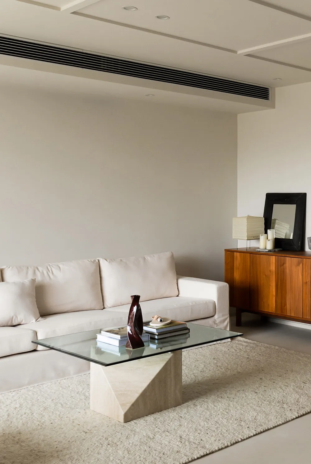 A modern living room with minimalist furniture and neutral-toned decor, featuring a glass-top coffee table, a wooden sideboar