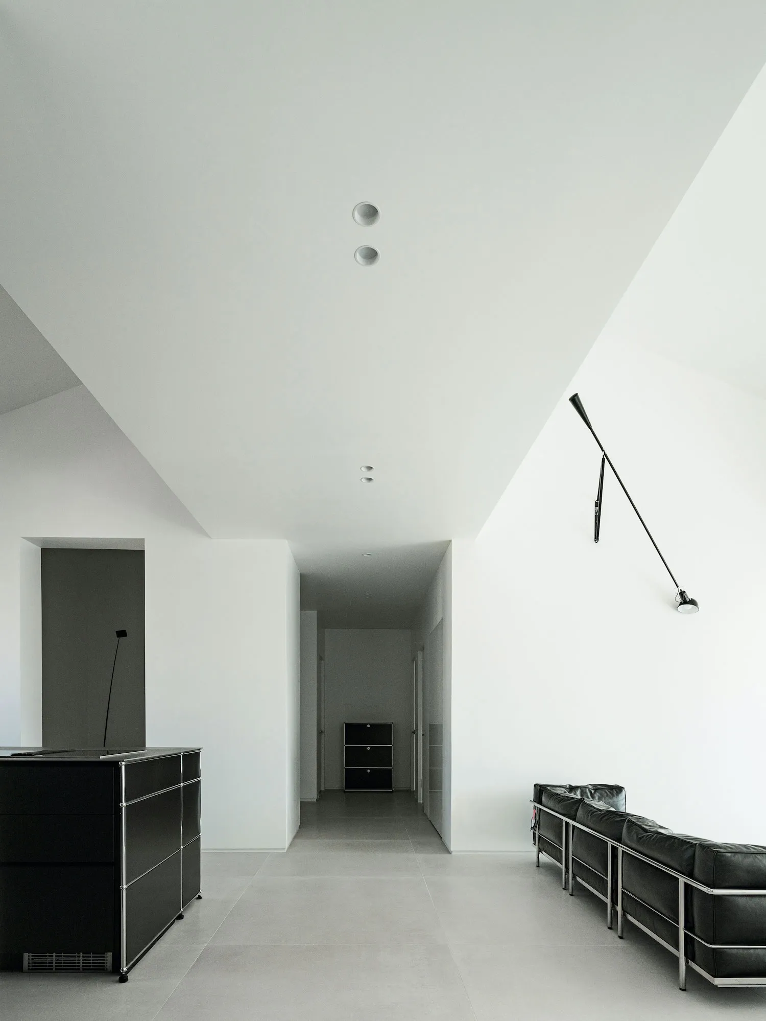 A minimalist interior space featuring a concrete floor with a long, sleek, black floor lamp and a black sofa with metal legs.