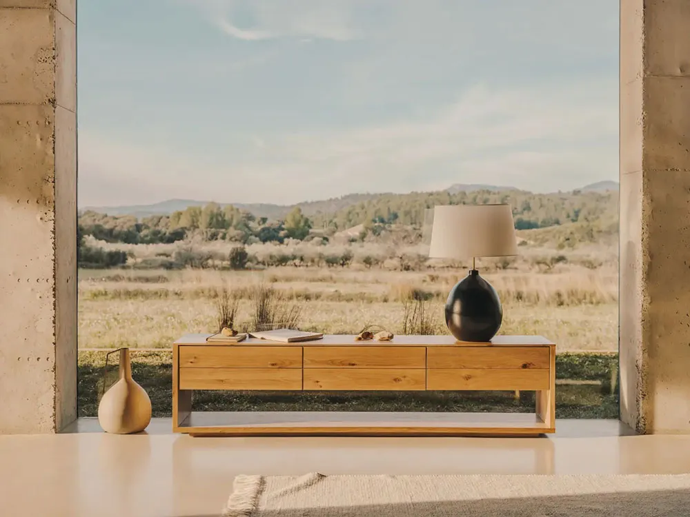 Minimalist interior design featuring wooden furniture, a lamp, and a vase in a spacious, open-concept room with a view of hil