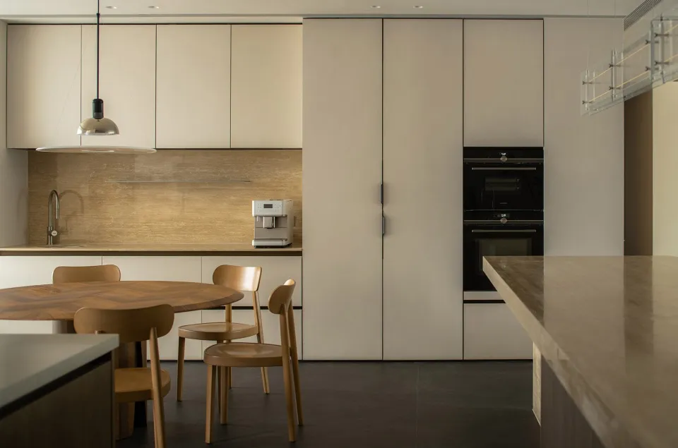 A modern kitchen with minimalist design featuring concrete countertops, wooden chairs, and stainless steel appliances.
