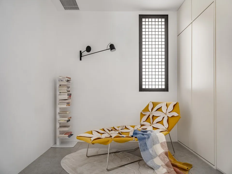 A minimalist bedroom featuring a yellow chaise lounge with flower upholstery, a blue and white striped blanket, and a bookshe