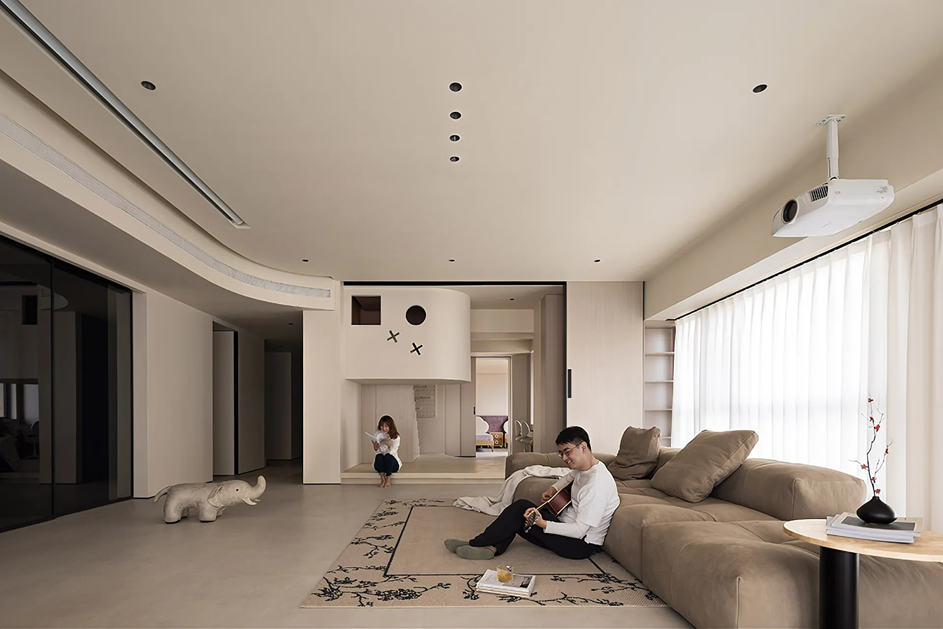 A modern minimalist living room with neutral-toned walls, furniture, and flooring, featuring a large sofa, a reading woman, a