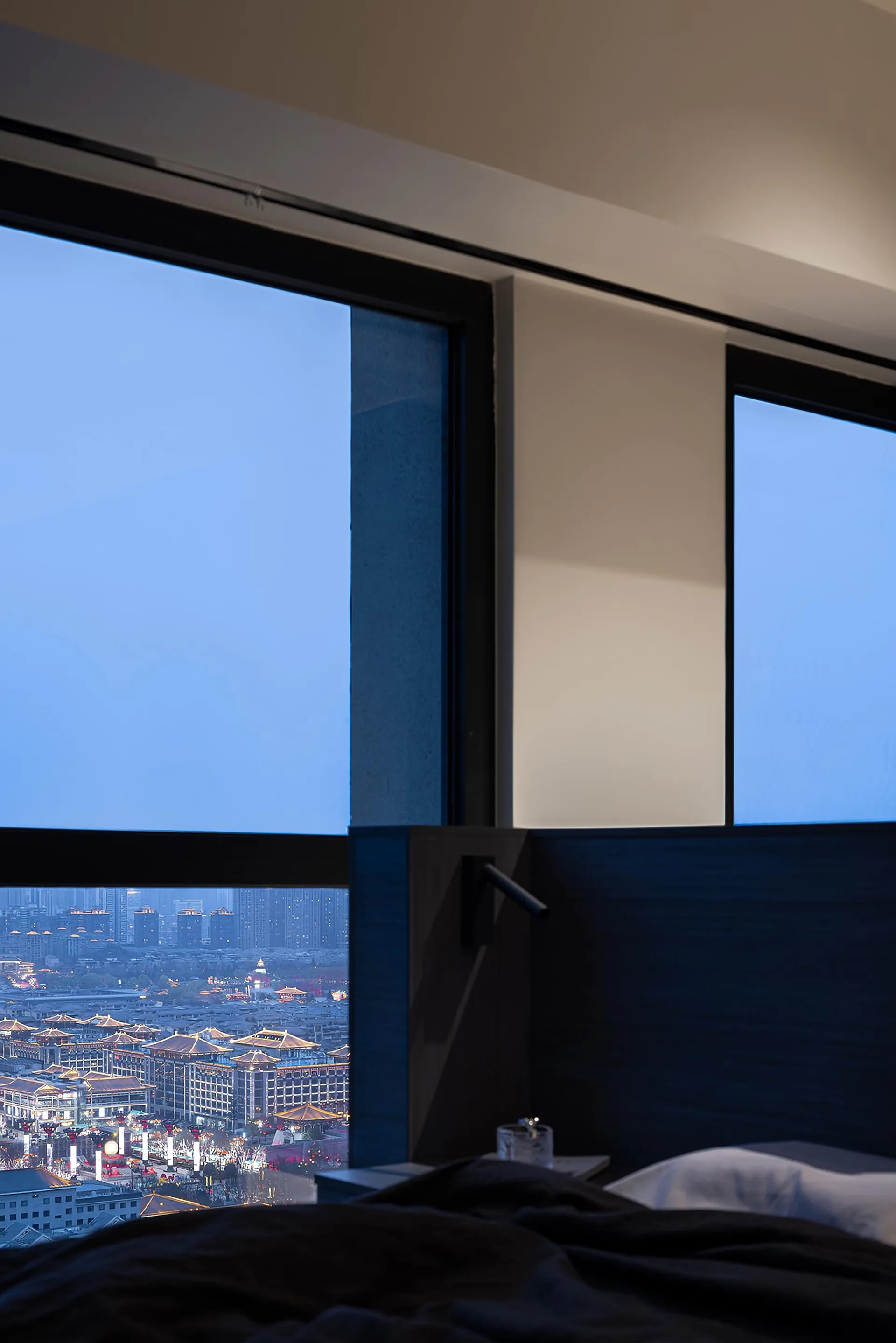 A modern bedroom featuring large glass windows overlooking a cityscape at dusk.