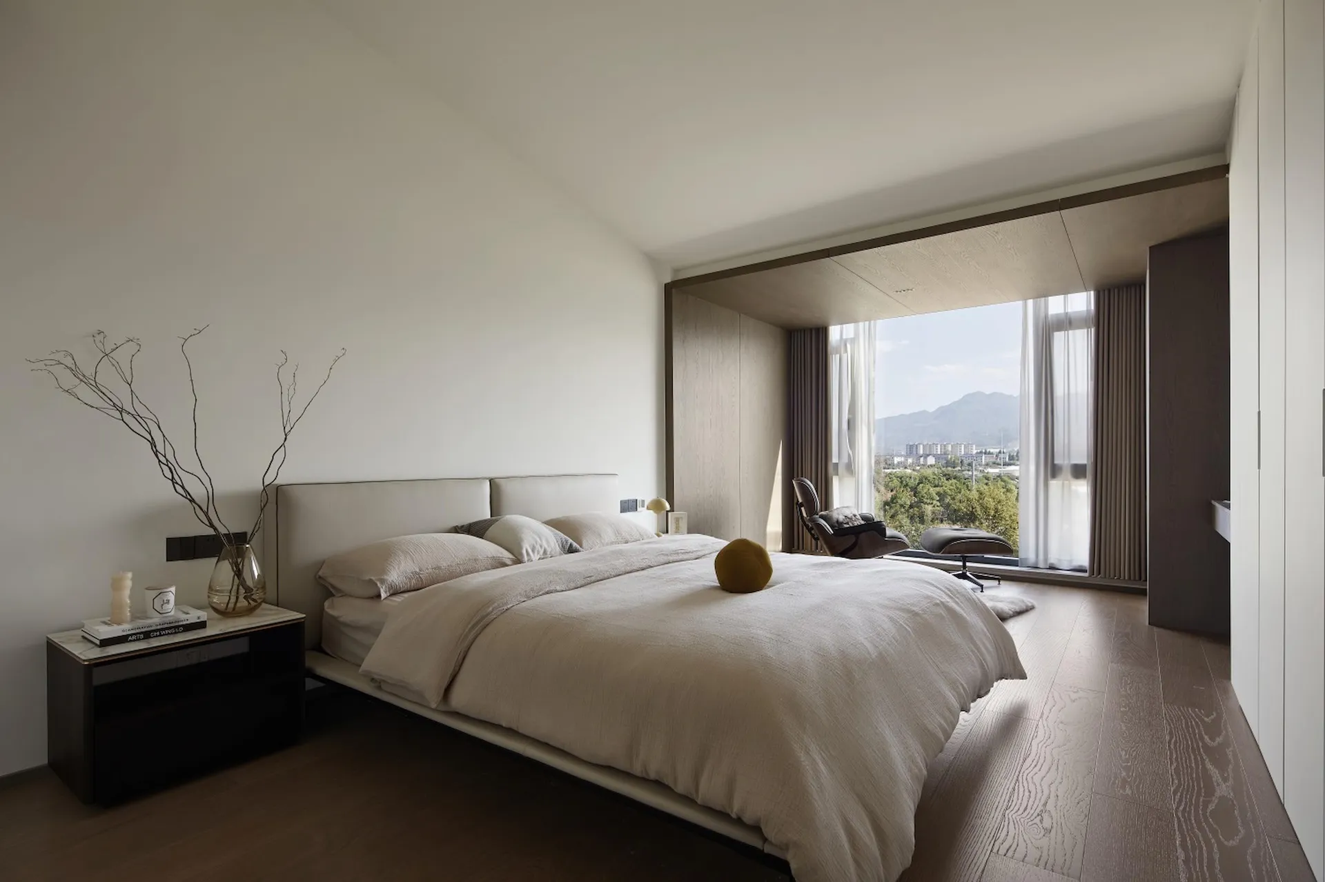 A modern minimalist bedroom featuring a wood floor, large window with mountain view, and simple furniture.