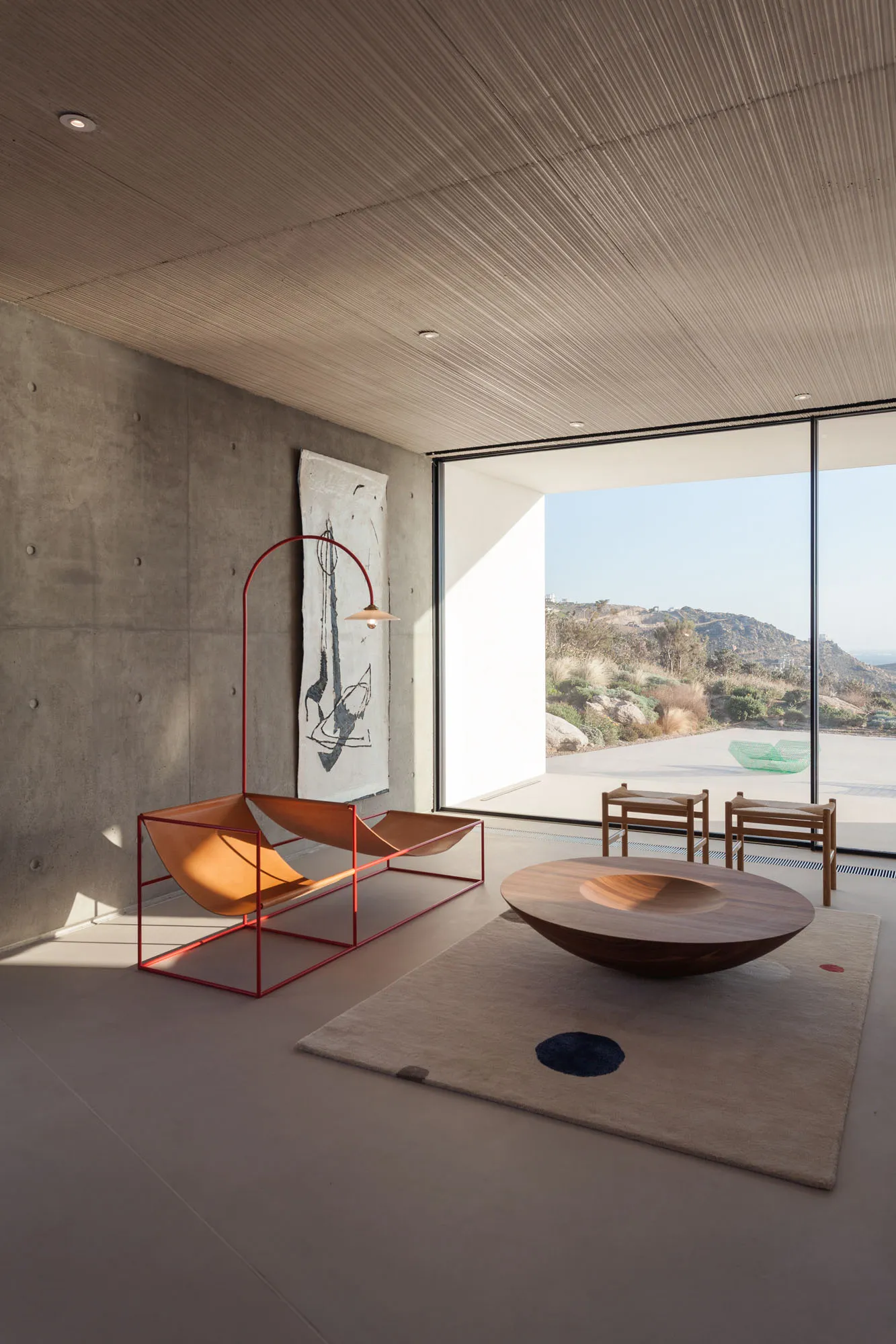 A spacious, minimalist living room featuring a concrete wall, wooden coffee table, and a large window offering a scenic view.