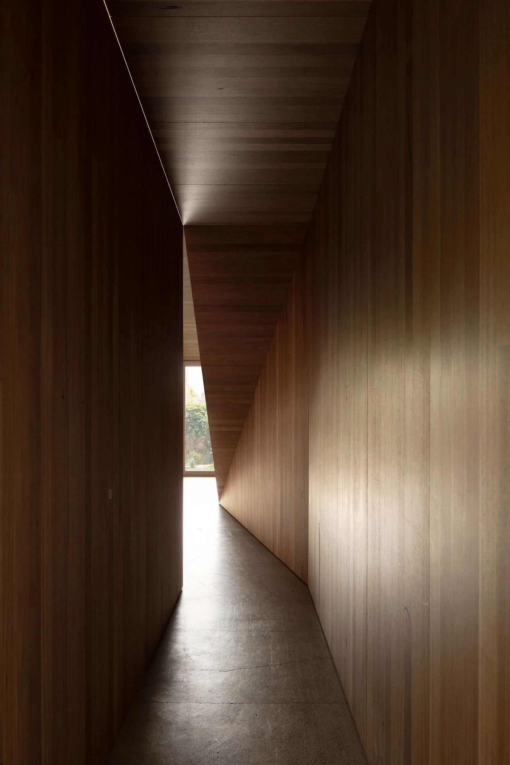 A narrow hallway with wooden walls, floor, and ceiling, featuring a window at the end.