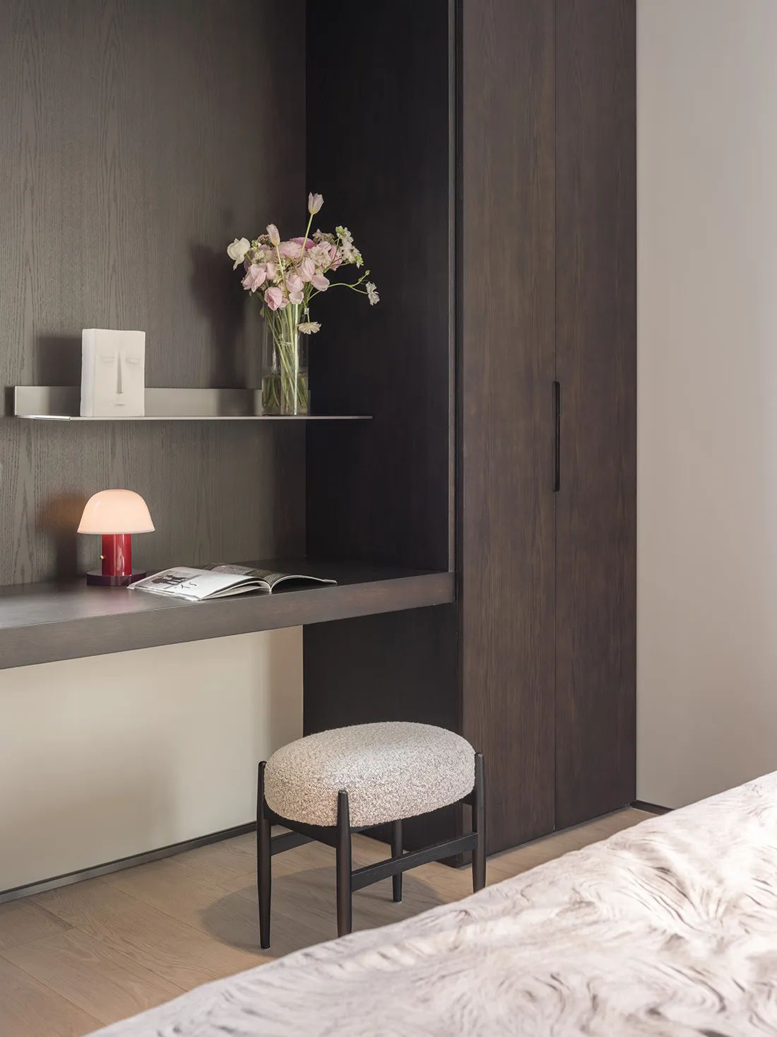 A minimalist bedroom featuring wooden furniture, a bed, a desk with a lamp and flowers, and a shelf.