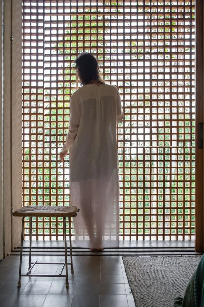 A woman is standing in front of a wooden lattice window with a stool in the foreground and a textured rug on the floor.