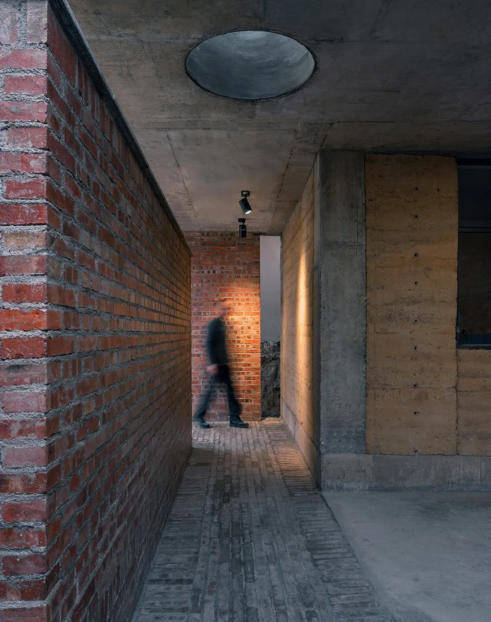 A minimalist corridor with brick and concrete walls, a concrete ceiling, and a person walking through a doorway.
