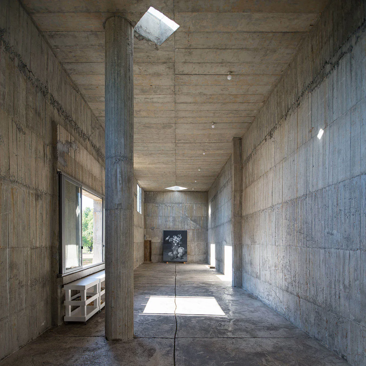 An empty minimalist gallery space with concrete walls and floor, featuring a single painting and a white shelf.