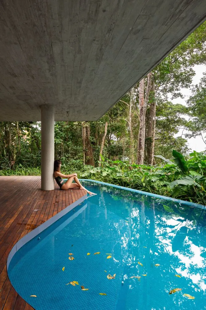 A woman sitting by a modern, concrete-covered pool surrounded by lush tropical vegetation.