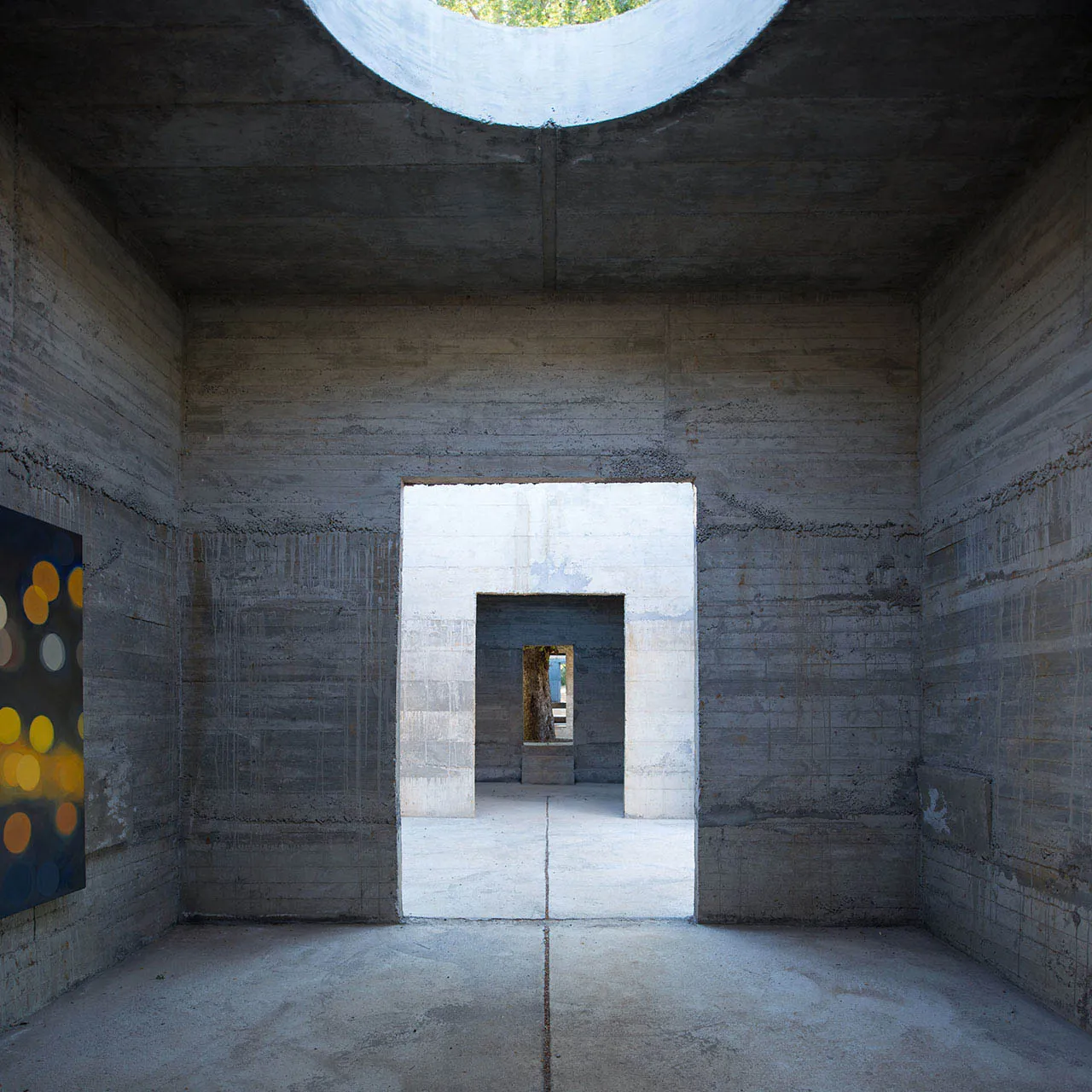 A minimalist interior space with concrete walls, a circular opening in the ceiling, and an open doorway leading to another ro