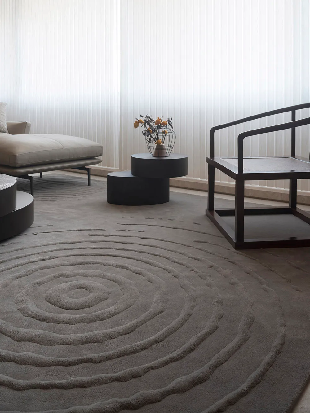 A minimalistic living room featuring a textured tiled rug, a white sofa, a black wooden armchair, and a side table with dried