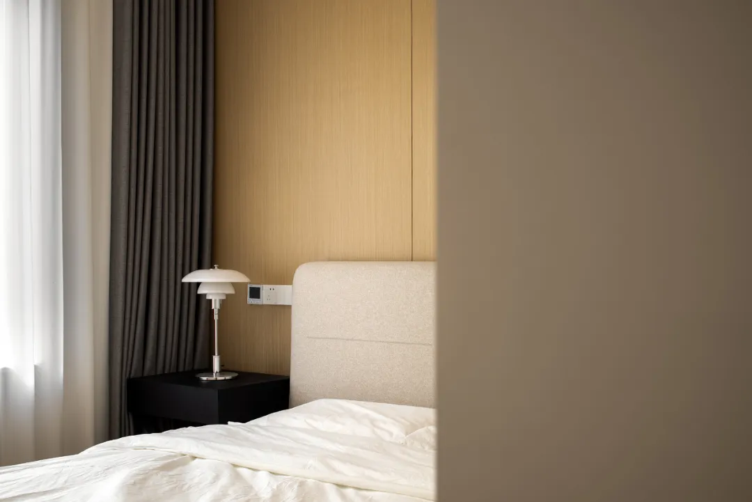 A minimalist bedroom with a white bed, grey curtains, a black side table with a white lamp, and a wooden headboard.