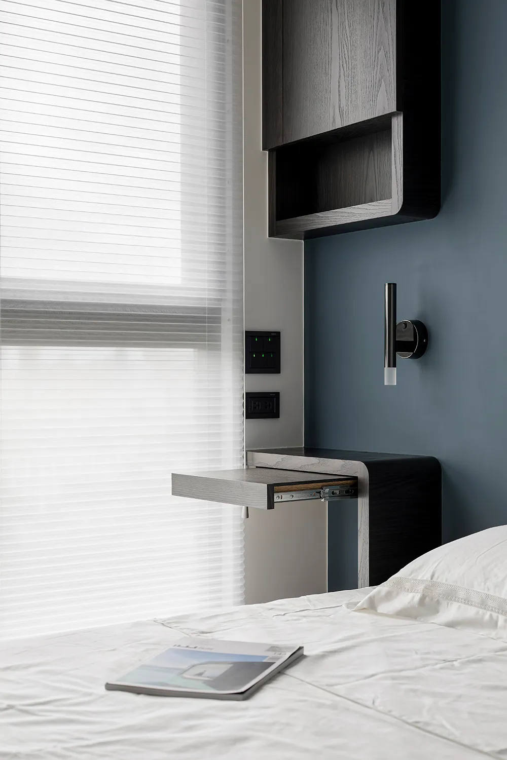 A minimalist bedroom featuring a wooden desk, blinds, and a comfortable bed with white bedding.