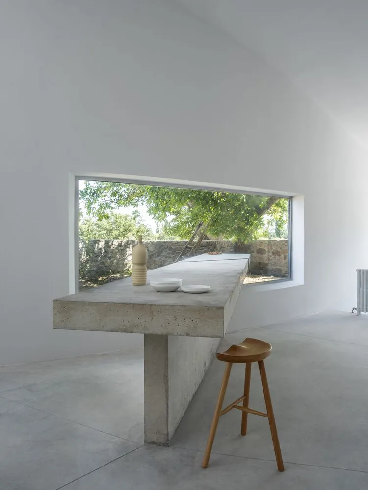 A minimalist concrete table with a window displaying greenery, a stool, and a vase.