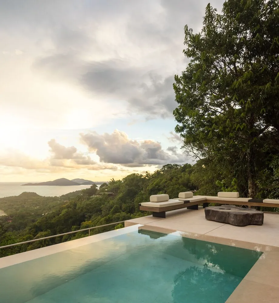 Modern outdoor living space featuring a concrete and glass infinity pool with a scenic view of the ocean and forested hills.
