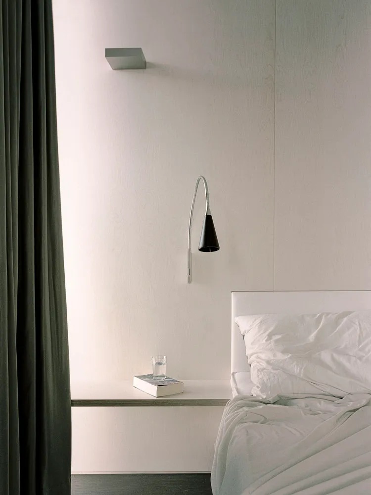 A minimalist bedroom with a white bed and grey curtains, featuring a white nightstand, a fabric lampshade, and a glass water