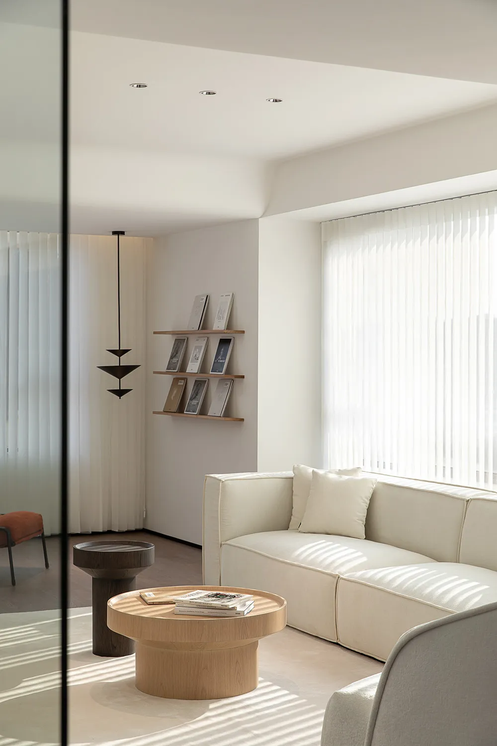 A minimalist living room featuring a light wood sofa and coffee table, with white curtains and a suspended light fixture.