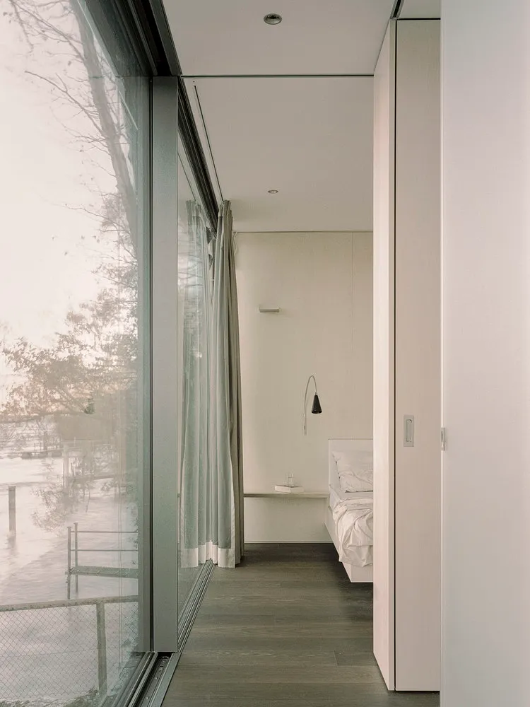 Modern minimalist bedroom with glass window, white walls, and wooden floor.