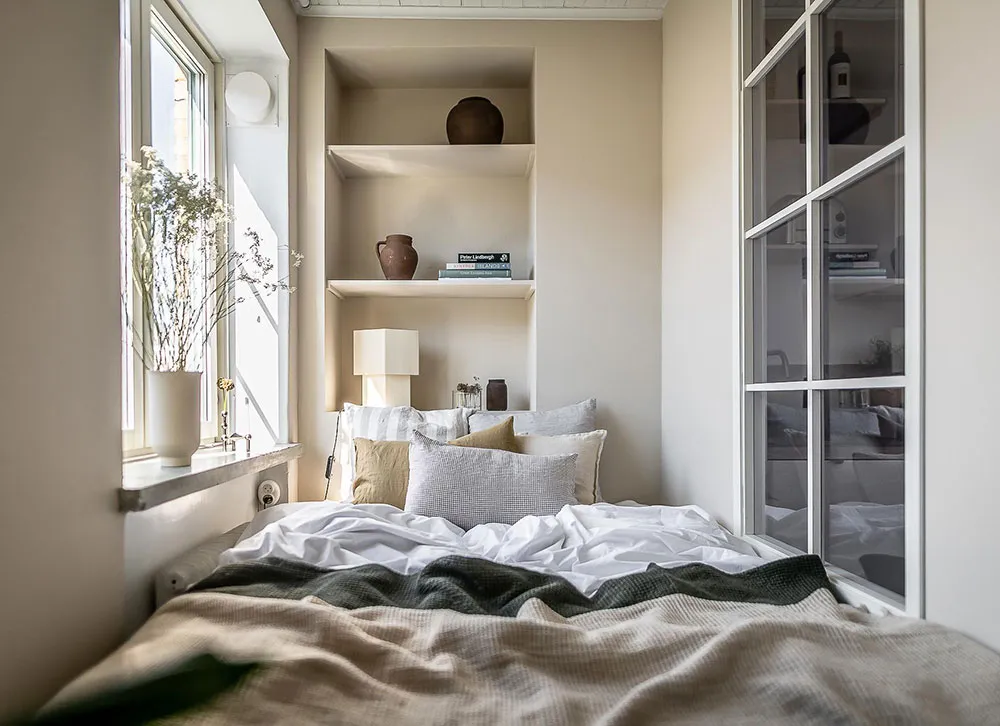 A cozy, minimalist bedroom with neutral-colored bedding, shelves filled with books and vases, and a window letting in natural