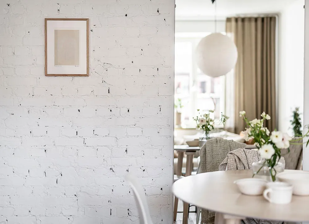 White brick wall in a minimalist dining room setting with a round wooden table, chairs, and decorative flowers.