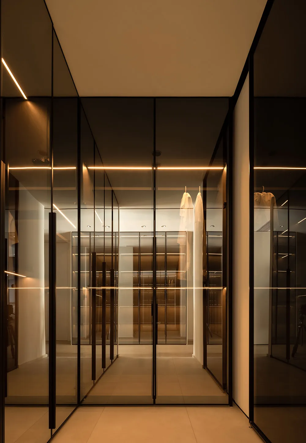 A modern clothes closet with glass walls and mirrors inside, illuminated by soft lighting.