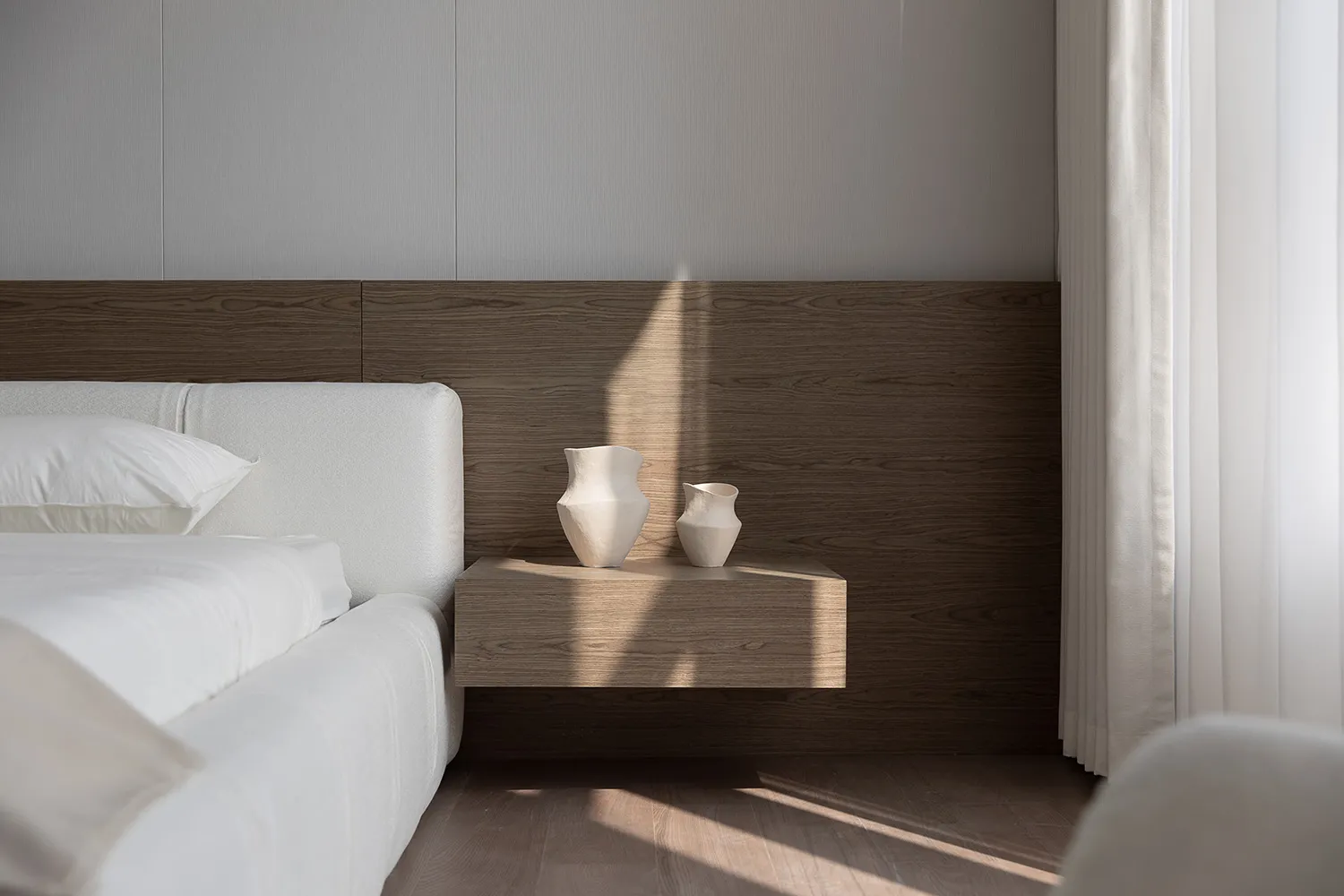 Minimalist bedroom featuring a white bed with fluffy pillows and a floating wooden nightstand with two decorative vases.