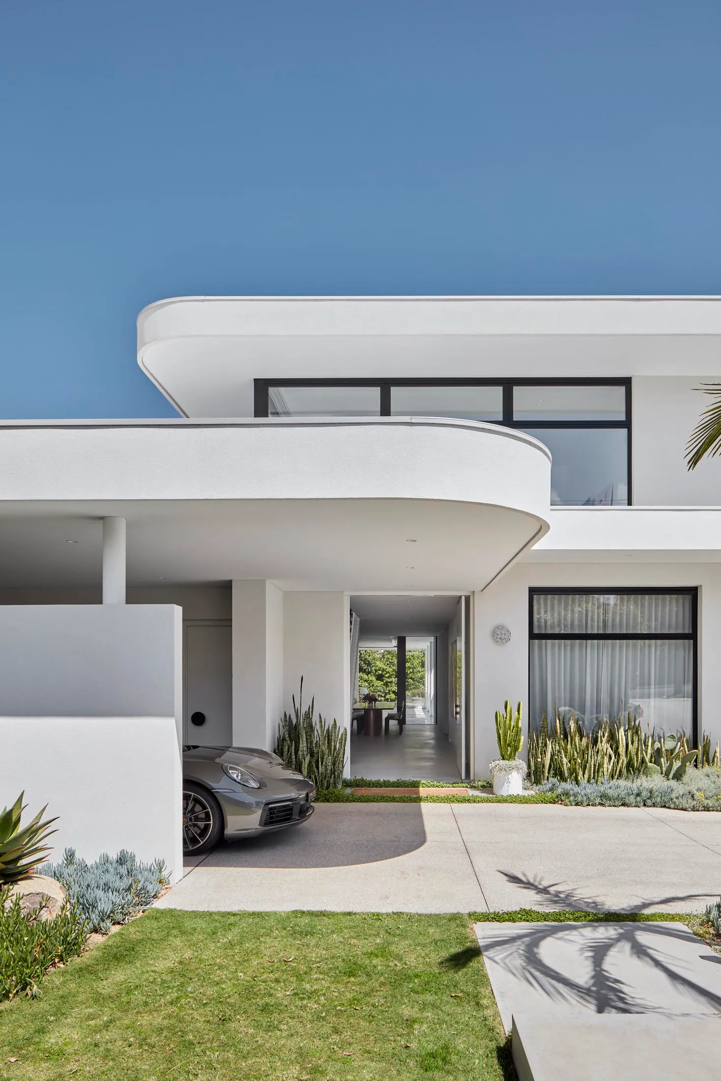 A modern house with a white, curved concrete exterior, featuring large windows and a driveway with a parked car.