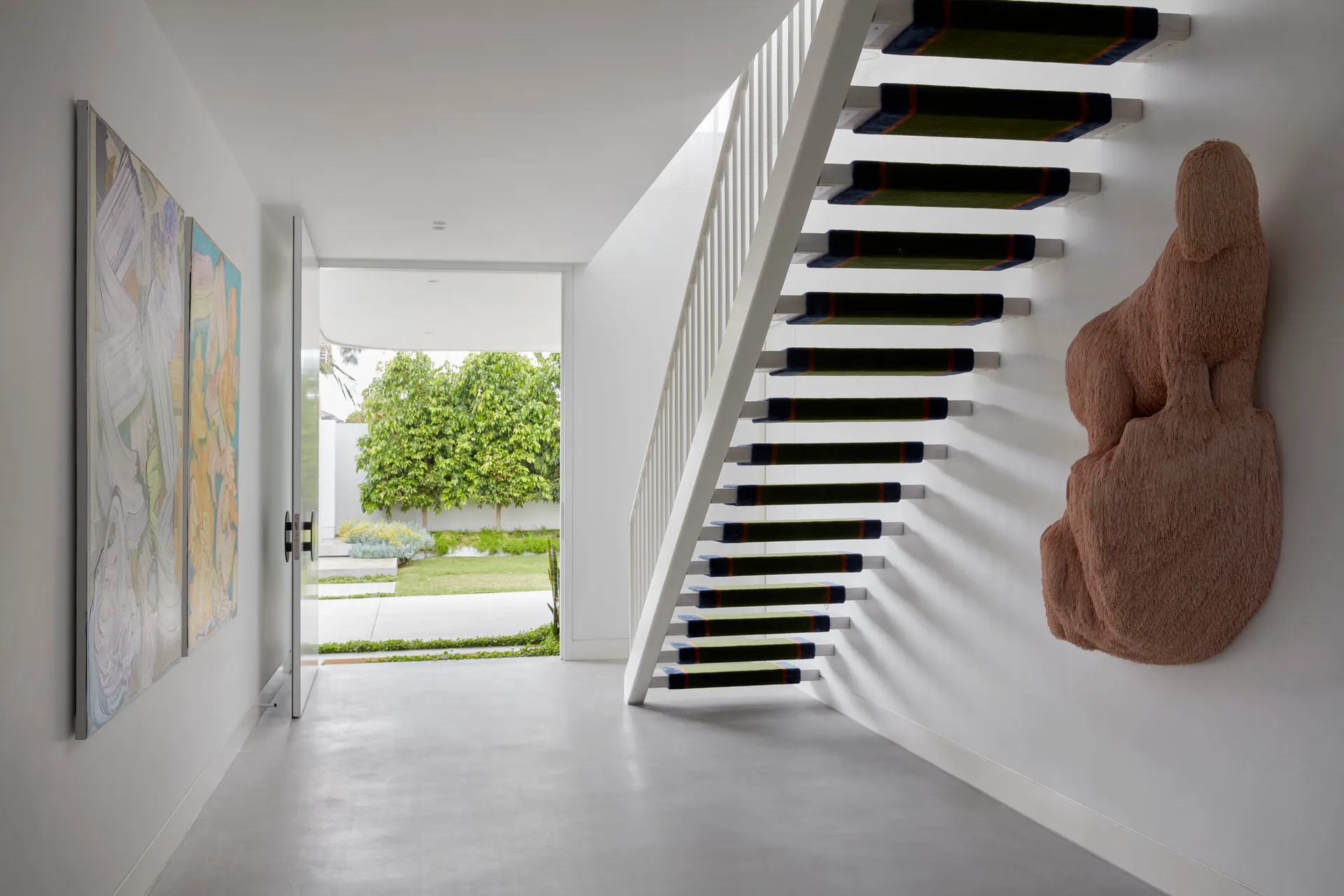A modern staircase with metal railings and green carpeted steps leading up to a white wall with a large, abstract sculpture m