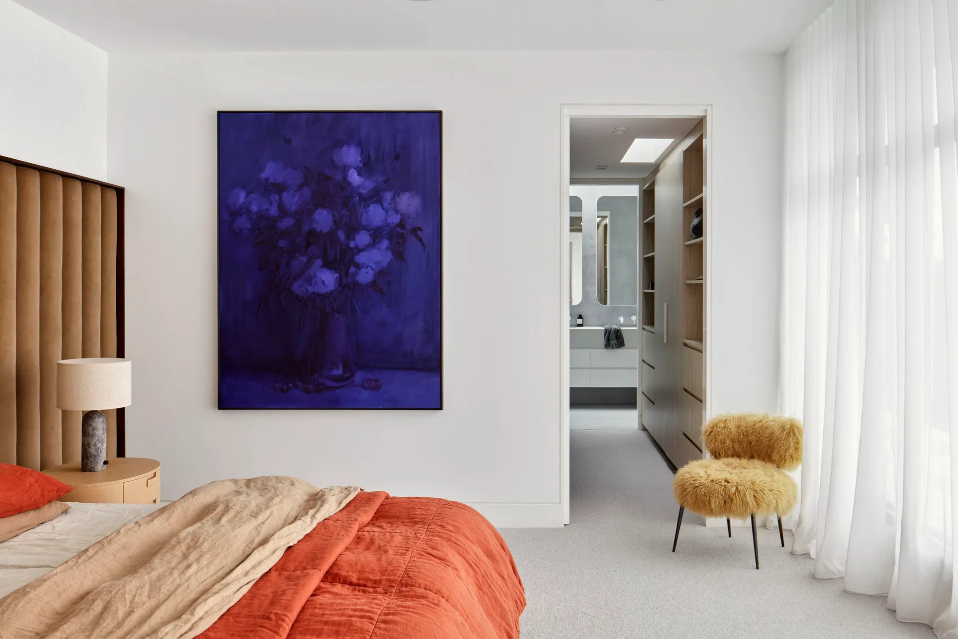 A minimalist bedroom featuring a bed with textured textiles, a fur stool, and a purple floral painting on the wall, with acce