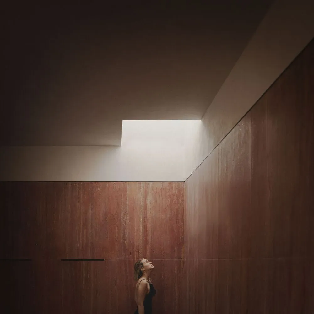 A minimalist interior designed with concrete materials, featuring a person looking upwards towards a skylight.