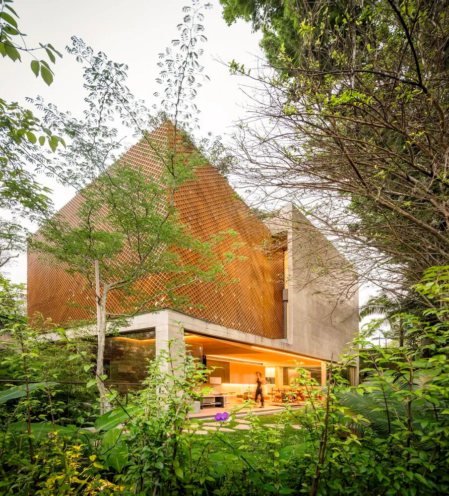 A modern house with a wooden textured exterior and a concrete base, surrounded by lush greenery and a person walking.