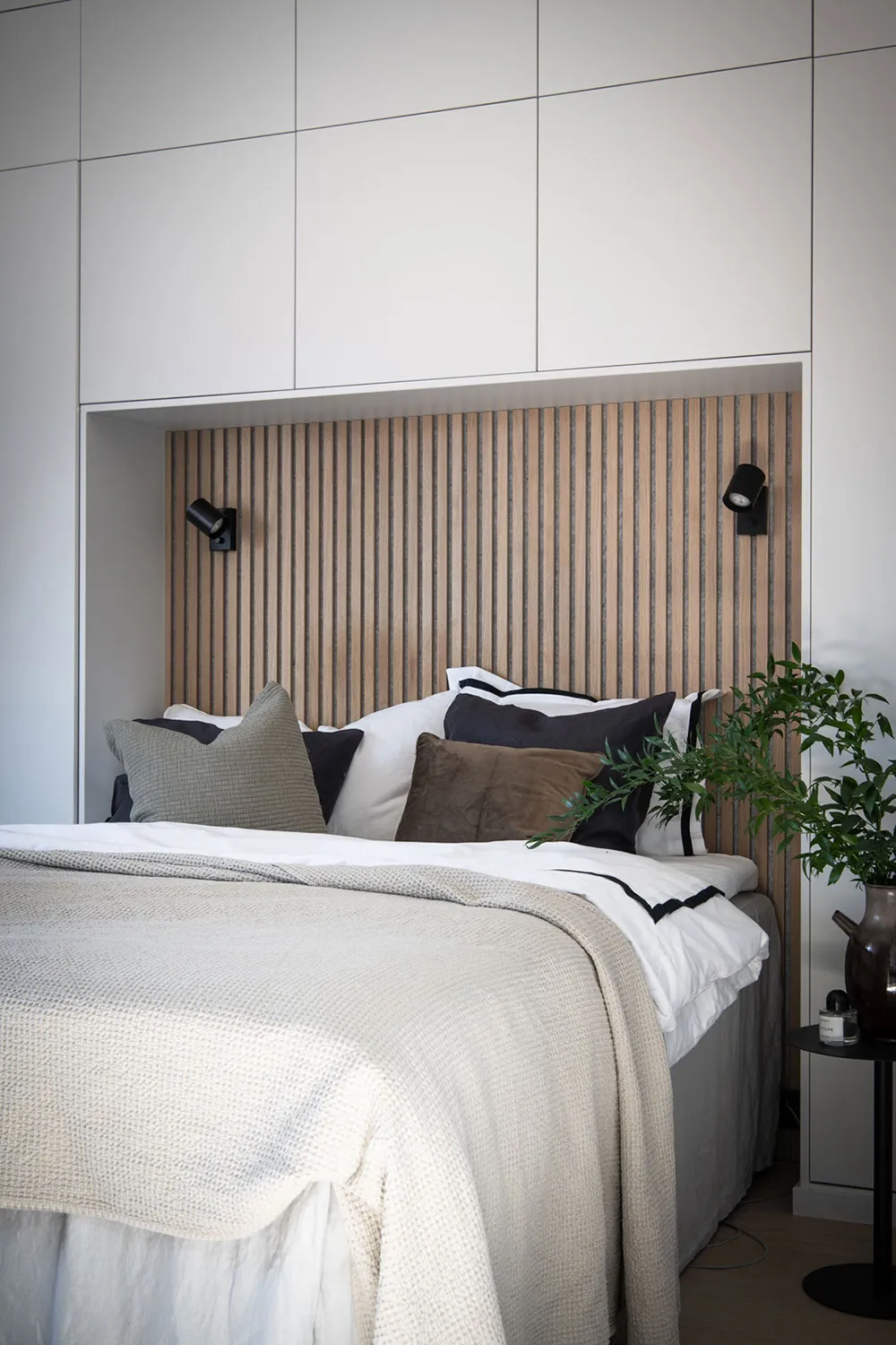 Modern minimalist bedroom interior with a wooden paneled headboard, white bedding, and a green plant on a side table.