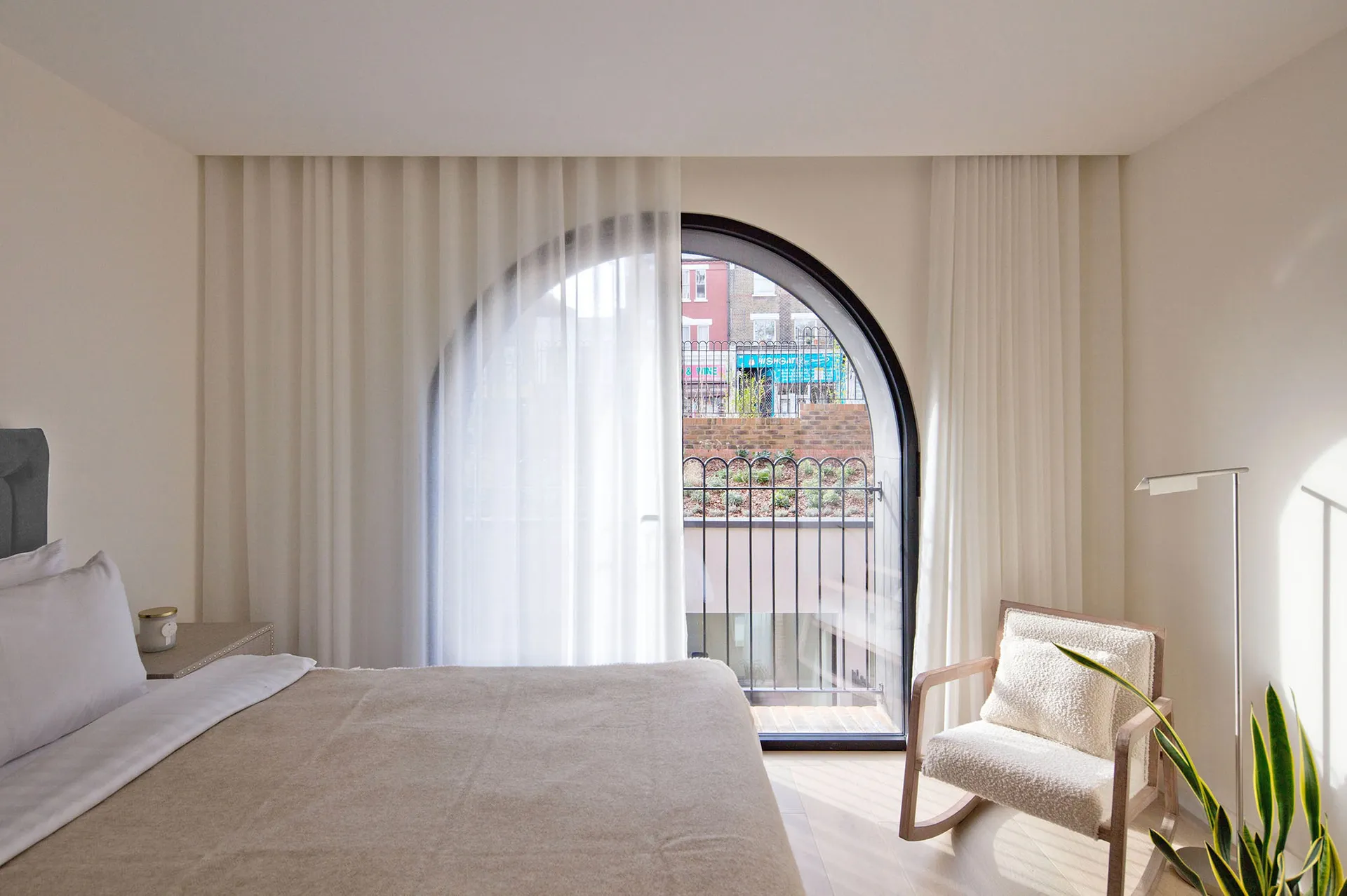 A minimalist bedroom with a large arched window, a bed with light-colored bedding, and a rocking chair.