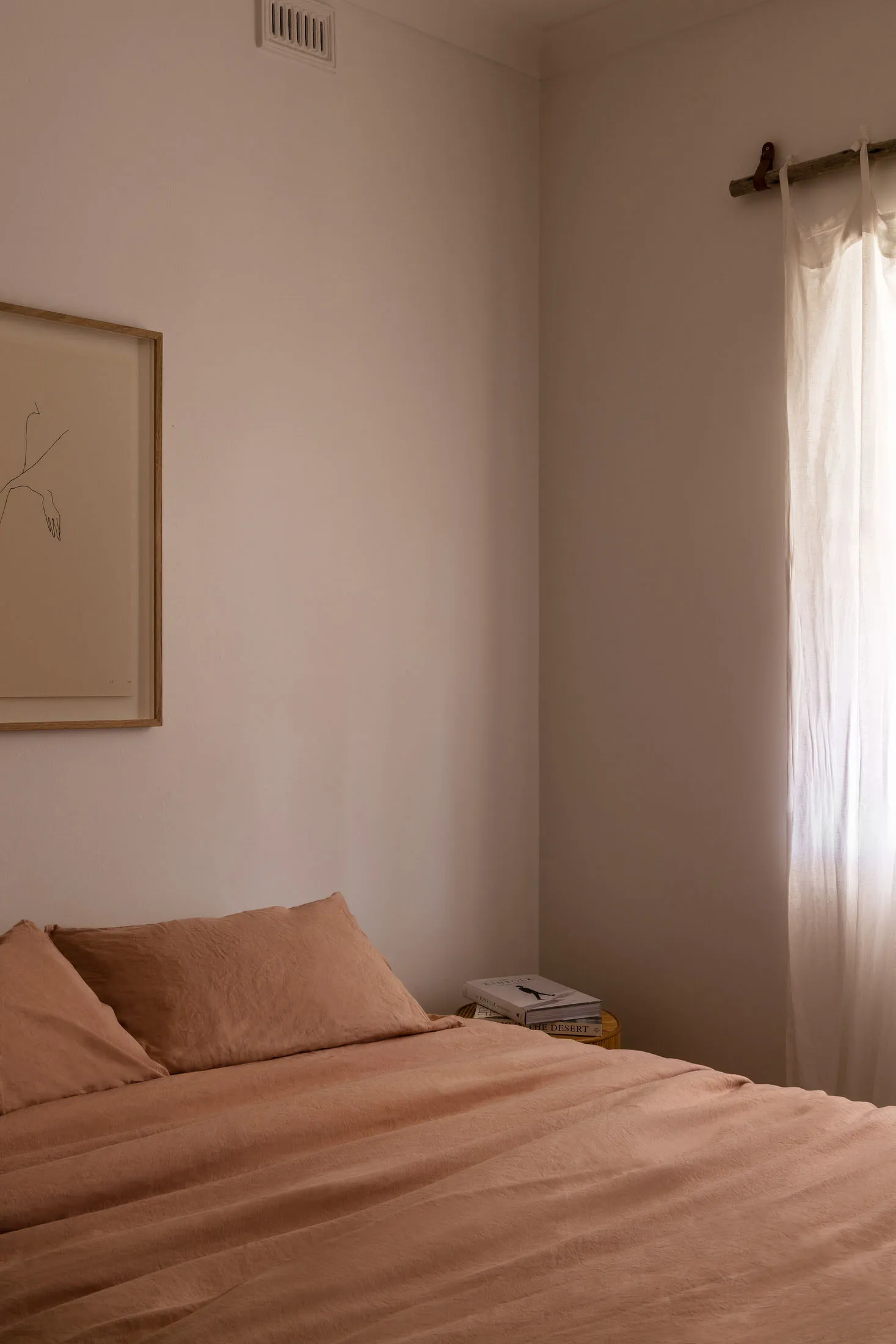 A minimalist bedroom with light brown fabric bedding and a window with light filtering through the curtain.
