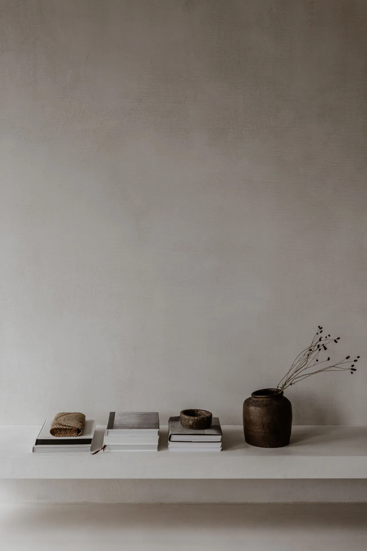 A minimalist shelf arranged with ceramic books and a vase containing greenery, against a textured wall.