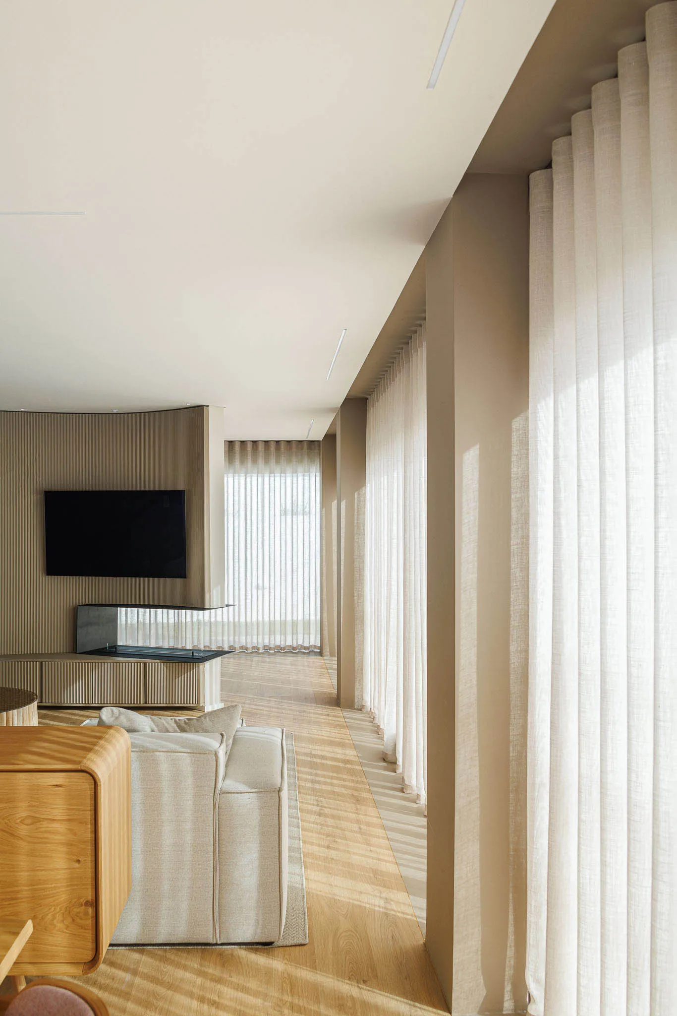 A modern minimalist living room featuring wooden flooring, cream fabric seating, and linen curtains.