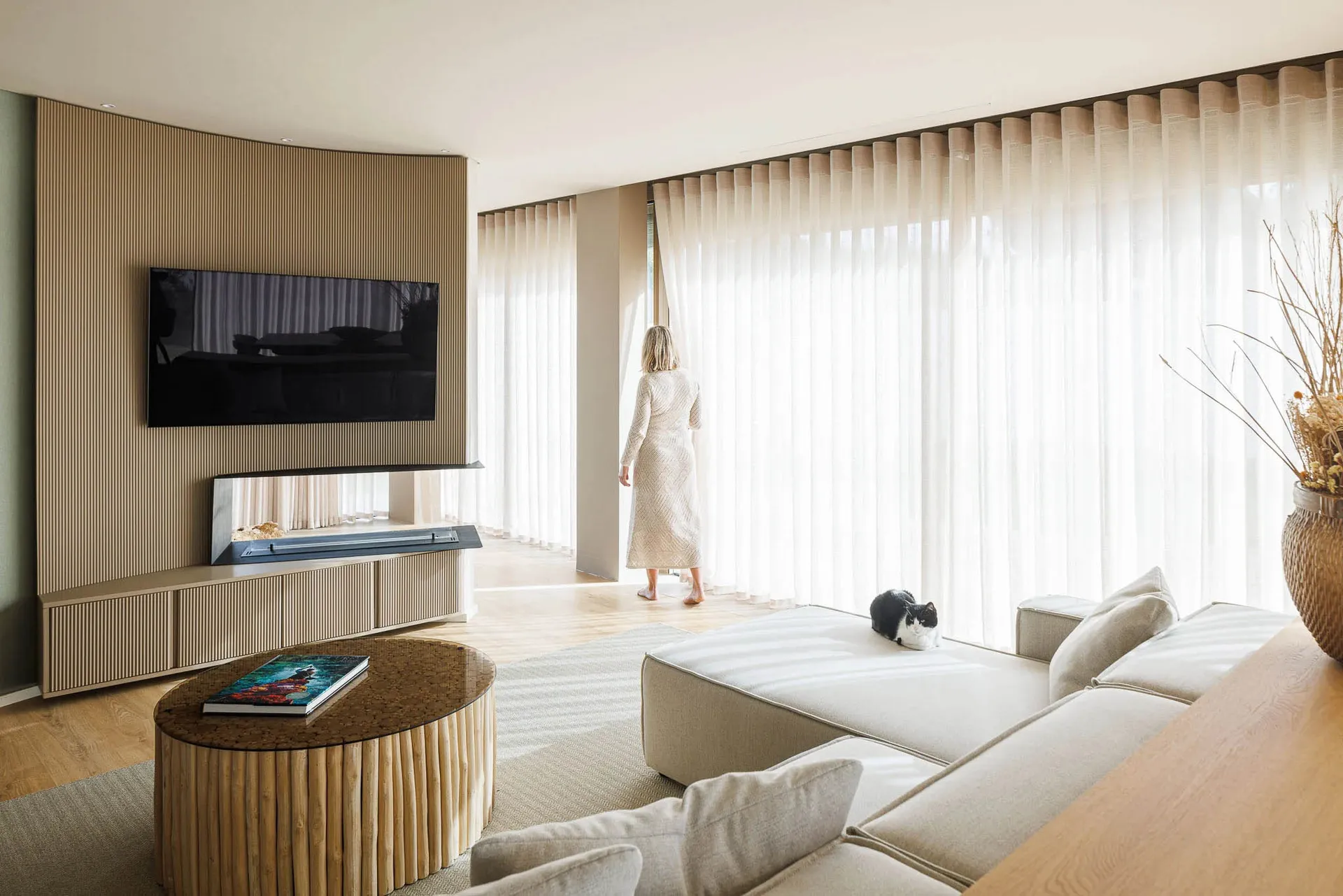 A modern living room with neutral tones and a cat resting on a sofa, as a woman stands by sheer curtains, looking out large w