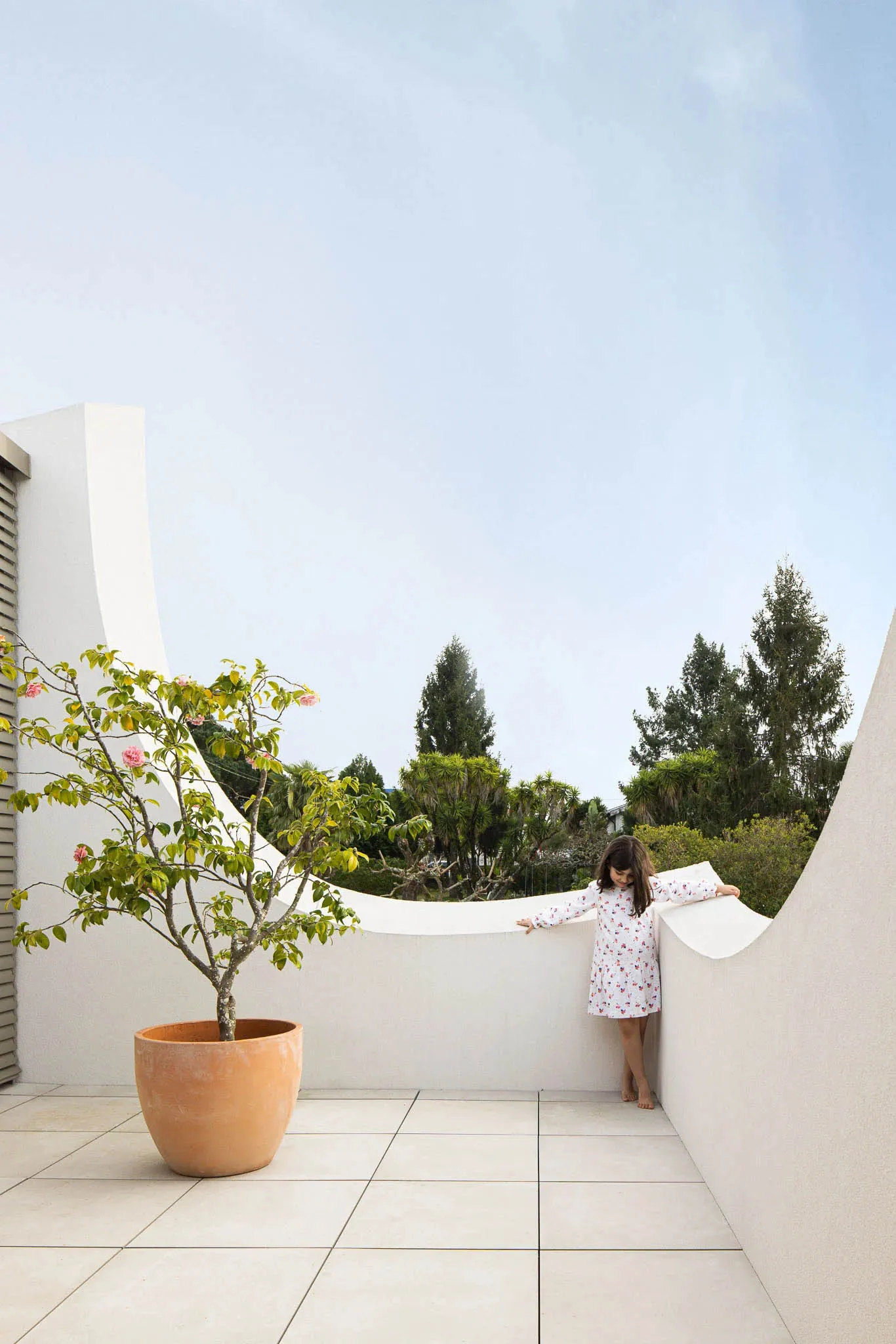 A modern rooftop garden featuring a child leaning against a curved white wall on a concrete floor, surrounded by greenery.