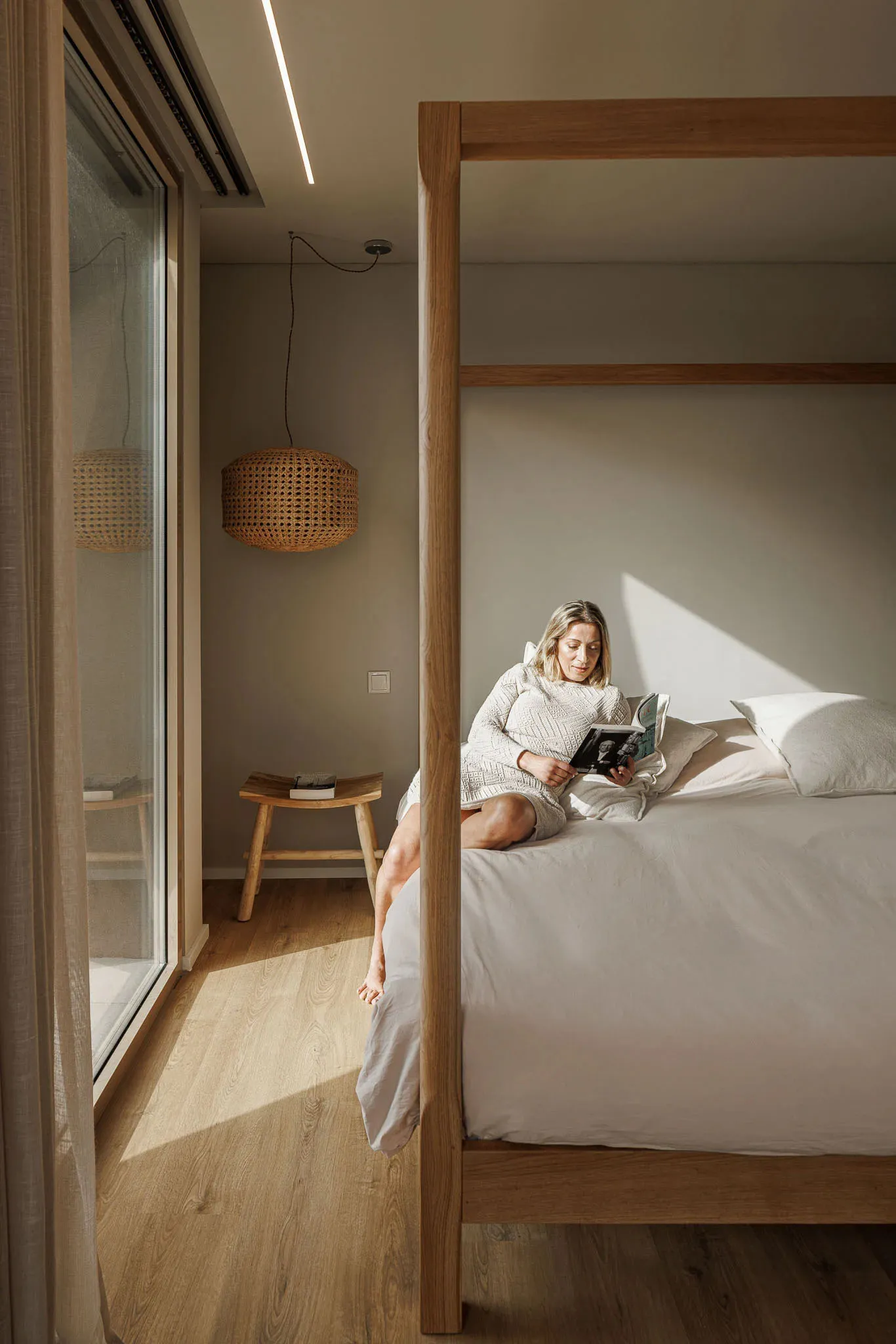 A woman in a minimalist bedroom reading a book while sitting on a wooden canopy bed with white linens and cushions, illuminat