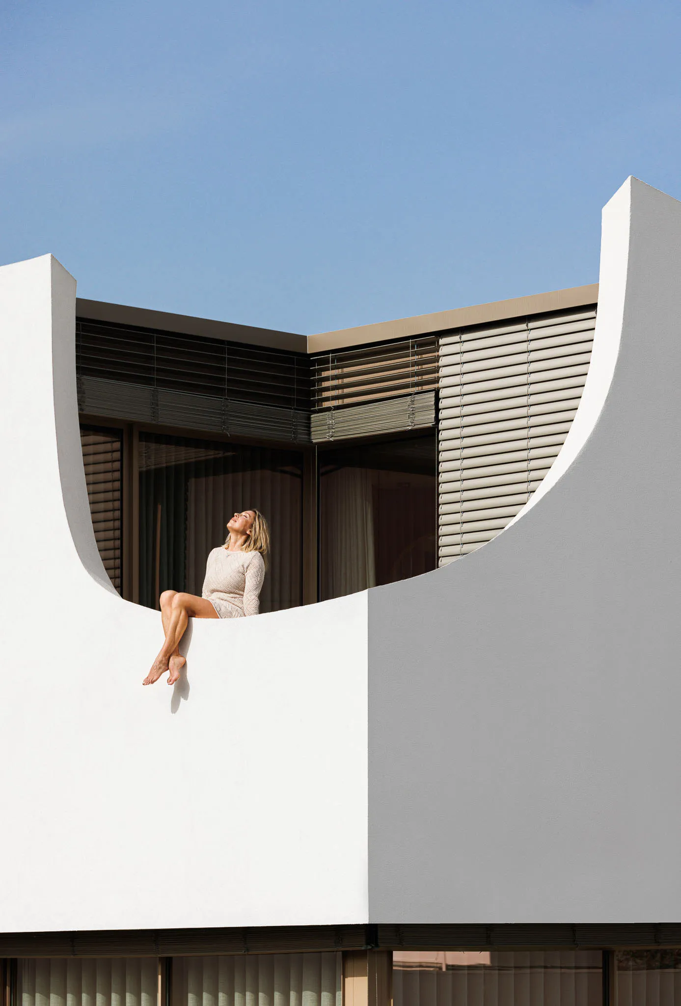 A woman sitting on a curved white architectural element against a clear blue sky.