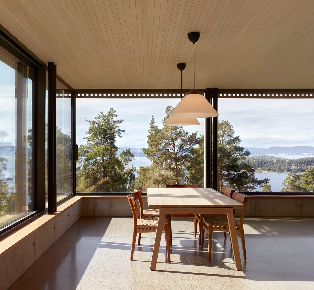 Contemporary dining room with a wooden table, chairs, and pendant lights, featuring a large window overlooking a forest and l