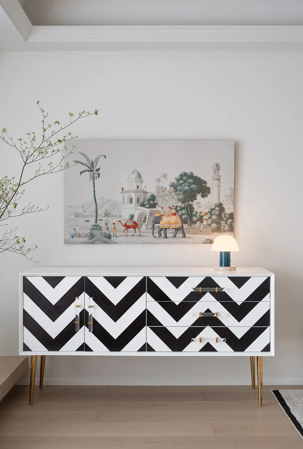 A modern living room with a white and black zigzag-patterned dresser, a table lamp, and a framed painting depicting a desert