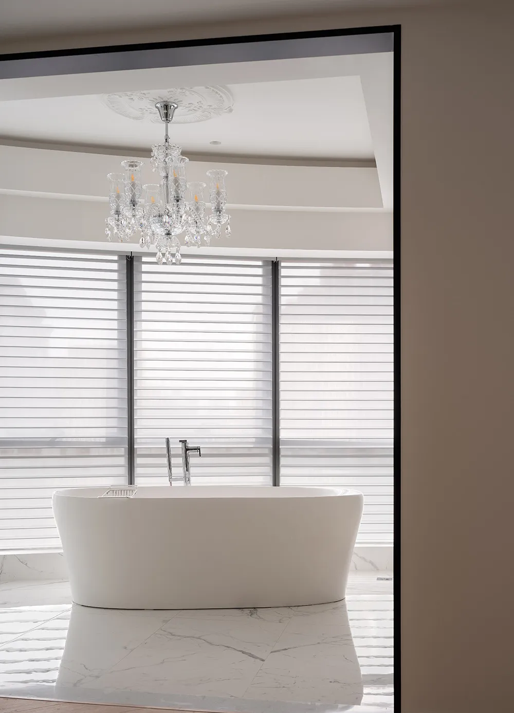A modern bathroom with a freestanding bathtub made of white marble, featuring a crystal chandelier and blinds in front of the
